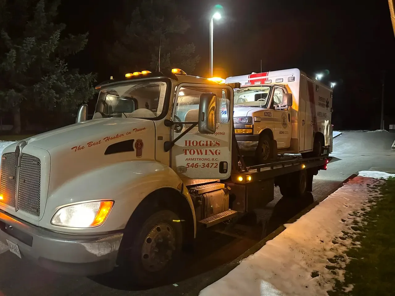 Hogie's white Kenworth deck truck hauling an ambulance at night, snow along the road